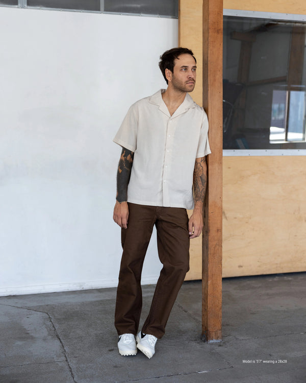Man wearing a beige shirt and brown pants standing against a white wall.