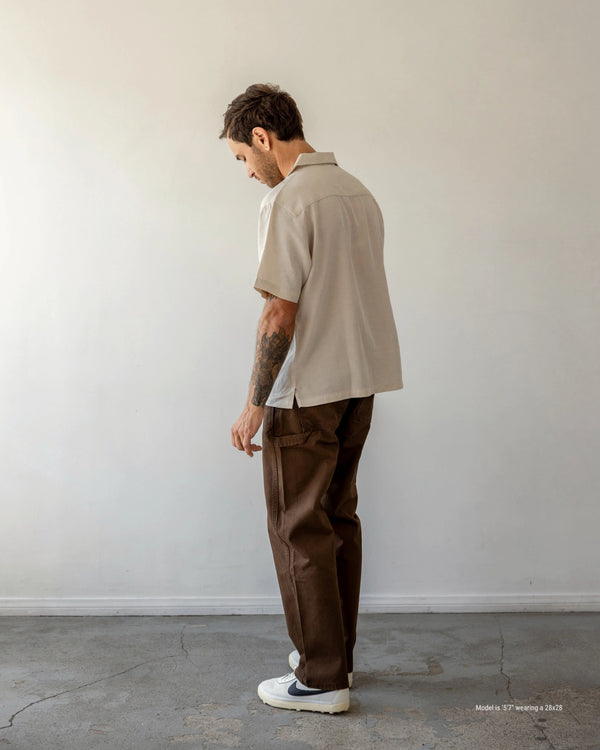 Man wearing a beige shirt and brown pants standing against a white wall.