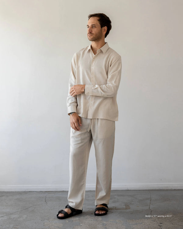 Man wearing a beige shirt and pants standing against a white wall.