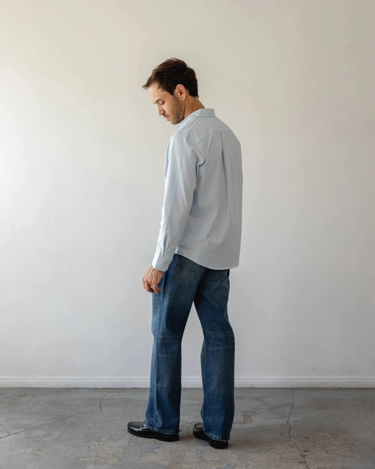 Man wearing a light blue shirt and dark jeans standing against a white wall.