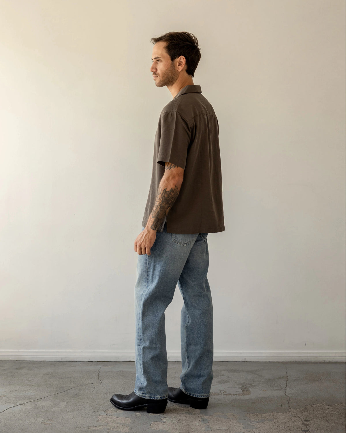 Shorter Man wearing a brown shirt and blue jeans standing against a plain white wall.