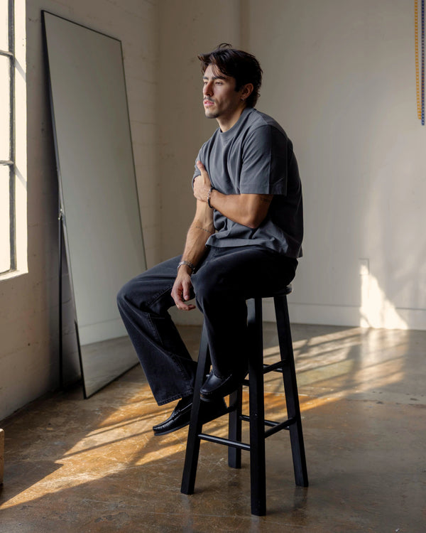 Shorter Man sitting on a stool in a room with a large mirror and sunlight streaming in.