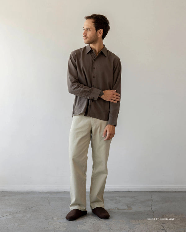 Man wearing a brown shirt and beige pants standing against a white wall.