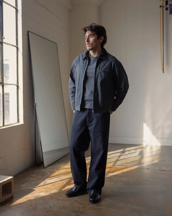 Short Man wearing a dark jacket and pants standing in a sunlit room with large windows.