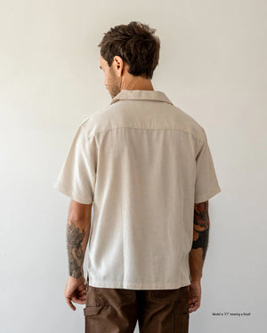 A short male model shown from the back wearing a natural Tumbled Linen Camp Shirt with brown trousers against a plain white studio background.