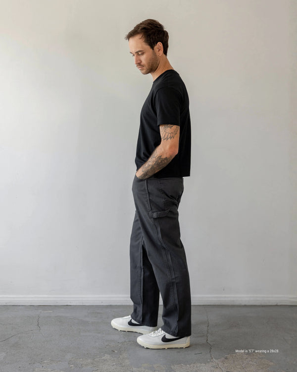 Man wearing a black t-shirt and dark pants standing against a plain white wall.