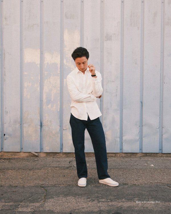 Shorter man wearing a white shirt and dark pants standing against a textured wall.