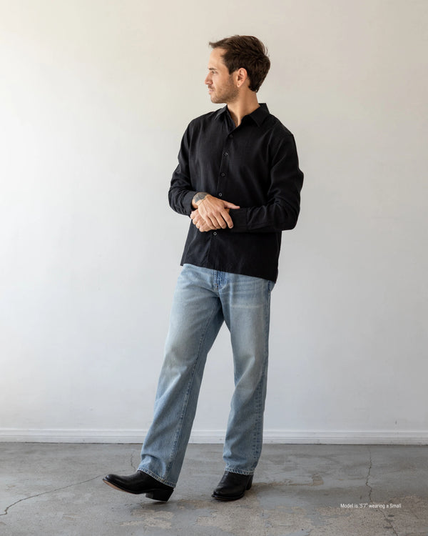 Man wearing a black shirt and light blue jeans standing against a white wall.
