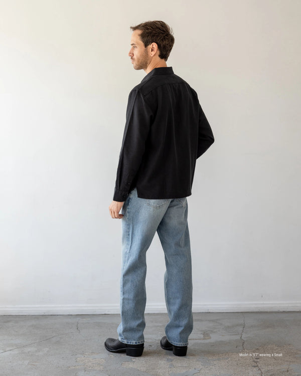 Man wearing a black shirt and light blue jeans standing against a white wall.