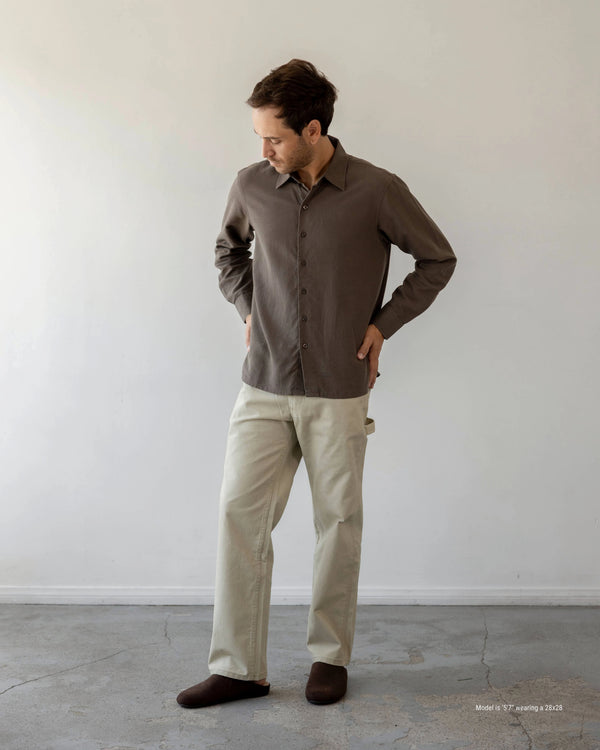 Shorter Man wearing a brown shirt and beige pants standing against a white wall.