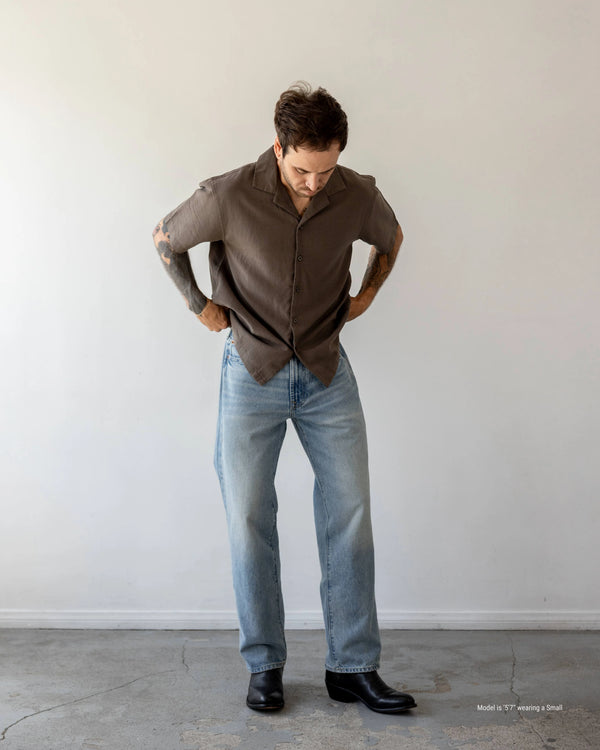 Man wearing a brown shirt and blue jeans against a white wall.
