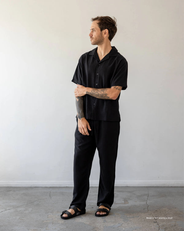 Man wearing a black outfit standing against a plain white wall.