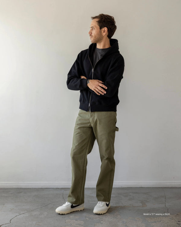 Man wearing a black jacket and green pants standing against a plain white wall.