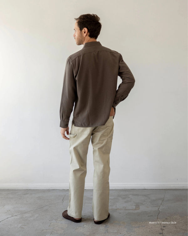 Man wearing a brown shirt and beige pants standing against a white wall.