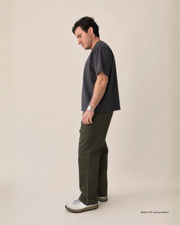 A man stands in profile wearing a dark charcoal gray short-sleeve T-shirt, olive green trousers, and white sneakers against a plain beige studio background.