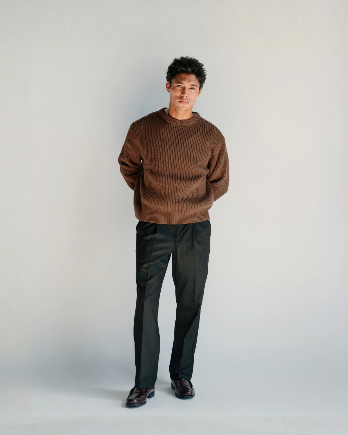 A short man wearing a brown ribbed knit sweater and dark charcoal trousers, standing against a light gray studio background.