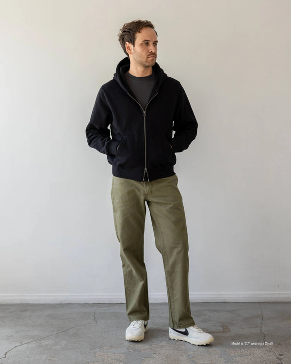 Shorter Man wearing a black jacket and green pants standing against a plain white wall.